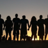 Group of people with linked hands silhouetted by sunset. 